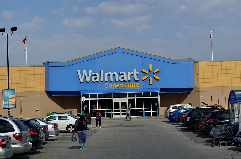 Exterior of a Walmart Supercentre store with blue and yellow signage, parked cars in the foreground, and shoppers walking toward the entrance.