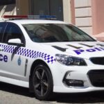White Western Australia Police–branded Holden Commodore VF II SV6 Sportwagon parked outside a historic sandstone building, showing its blue-and-white chequered livery and emergency lightbar.