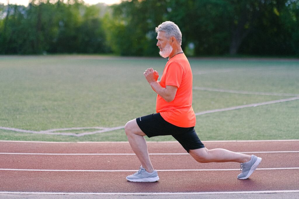 Senior man with gray hair and beard performing lunges on a running track, wearing bright orange t-shirt and black shorts.
