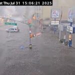 Floodwaters covering Northern Boulevard at Bell Boulevard in Bayside, Queens, with vehicles partially submerged and water flowing across the roadway.