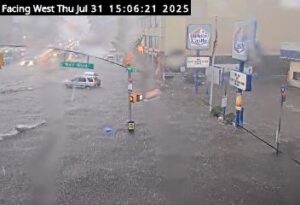 Floodwaters covering Northern Boulevard at Bell Boulevard in Bayside, Queens, with vehicles partially submerged and water flowing across the roadway.