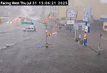 Floodwaters covering Northern Boulevard at Bell Boulevard in Bayside, Queens, with vehicles partially submerged and water flowing across the roadway.