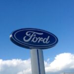 A tall Ford logo sign mounted on a pillar against a bright blue sky.