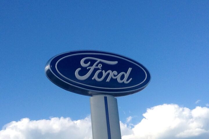 A tall Ford logo sign mounted on a pillar against a bright blue sky.