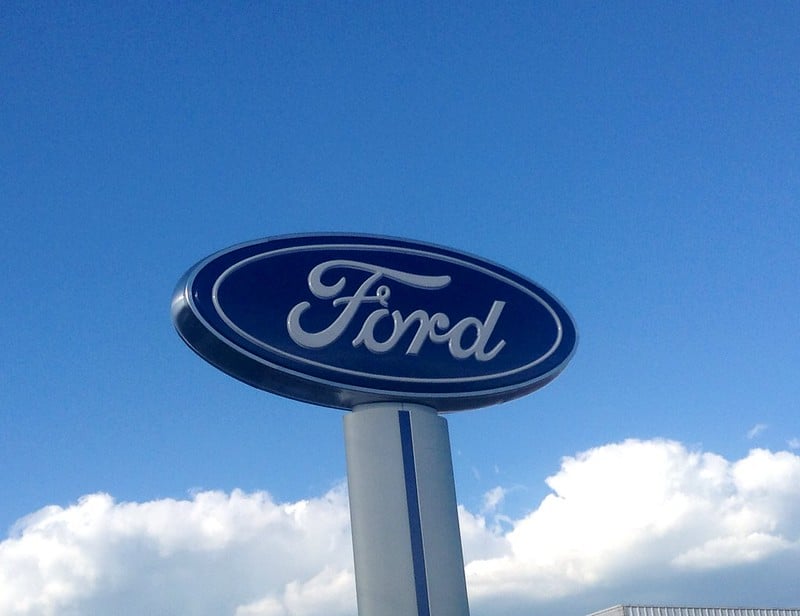A tall Ford logo sign mounted on a pillar against a bright blue sky.