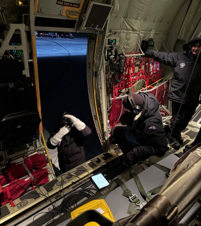 Personnel in heavy parkas stand at the open side ramp of a Hercules, preparing gear against a snowy backdrop.