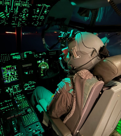 An RNZAF crew member in flight suit and helmet uses night-vision goggles inside the Hercules cockpit at night.