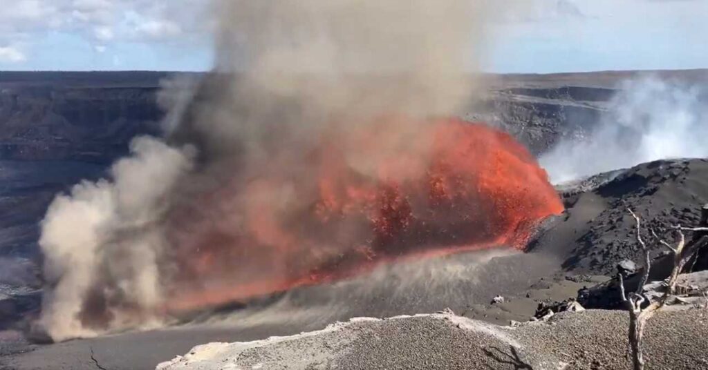 Glossy orange lava cascading at an angle from Kīlauea’s north vent, with glowing arcs of molten rock extending across the crater floor under a hazy plume.