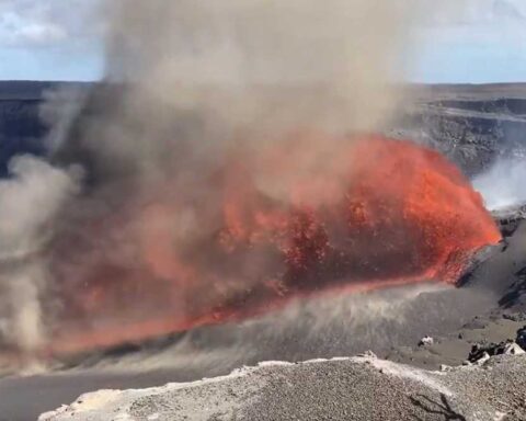 Glossy orange lava cascading at an angle from Kīlauea’s north vent, with glowing arcs of molten rock extending across the crater floor under a hazy plume.