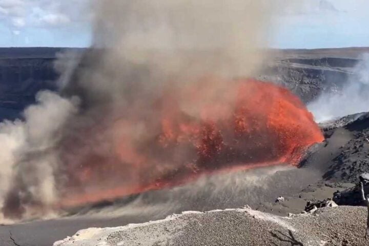 Glossy orange lava cascading at an angle from Kīlauea’s north vent, with glowing arcs of molten rock extending across the crater floor under a hazy plume.