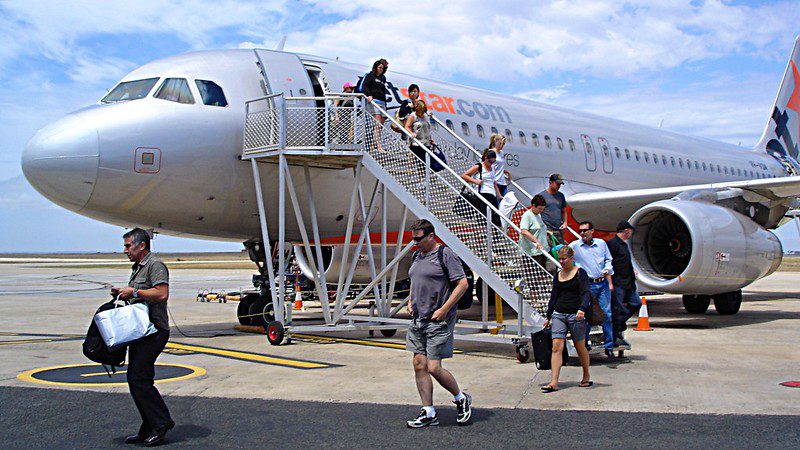 Passengers walking down aircraft stairs from Jetstar Airbus A320-200 plane at Avalon Airport with ground crew and luggage handling visible on tarmac
