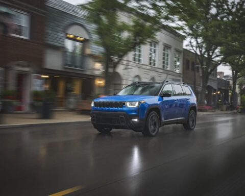 Blue 2026 Jeep Cherokee hybrid SUV driving on a wet city street, featuring the classic seven-slot grille and more upright, boxier design than its predecessor.