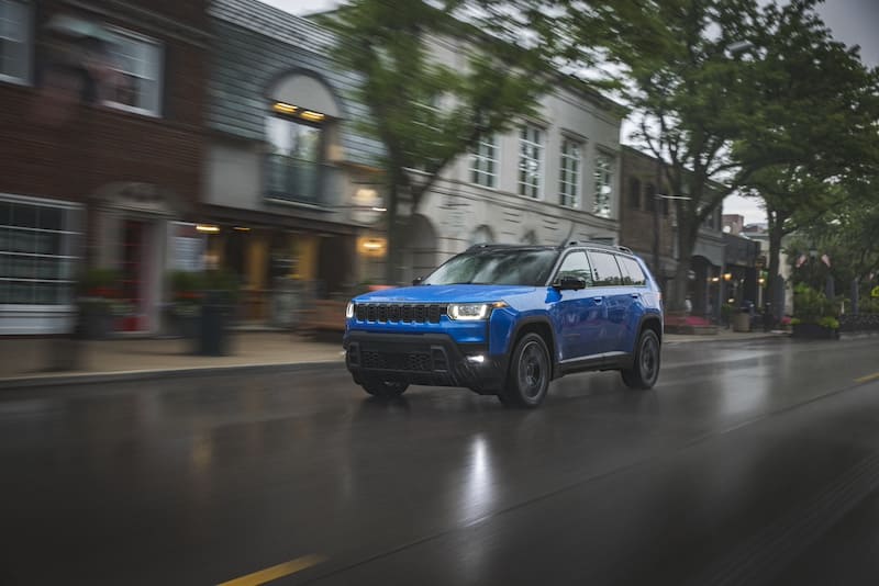 Blue 2026 Jeep Cherokee hybrid SUV driving on a wet city street, featuring the classic seven-slot grille and more upright, boxier design than its predecessor.