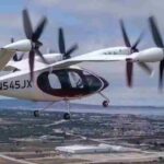 A Joby Aviation electric vertical takeoff and landing (eVTOL) aircraft in mid-flight over a coastal urban area, displaying the registration number N545JX.