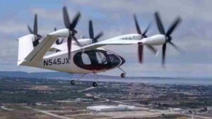 A Joby Aviation electric vertical takeoff and landing (eVTOL) aircraft in mid-flight over a coastal urban area, displaying the registration number N545JX.