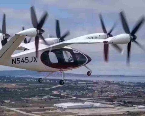 A Joby Aviation electric vertical takeoff and landing (eVTOL) aircraft in mid-flight over a coastal urban area, displaying the registration number N545JX.