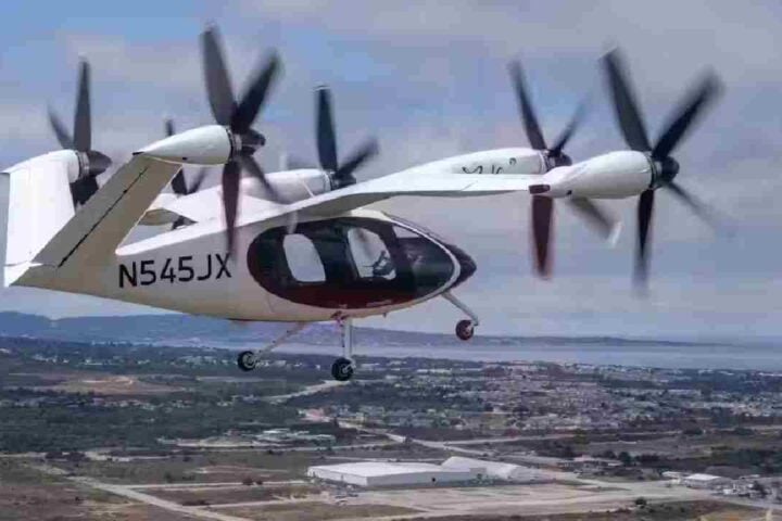 A Joby Aviation electric vertical takeoff and landing (eVTOL) aircraft in mid-flight over a coastal urban area, displaying the registration number N545JX.