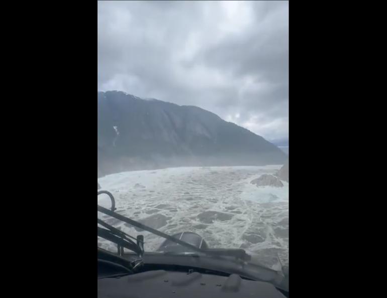 Aerial view of a fjord in Southeast Alaska filled with debris and rocks from a landslide, with mountains and cloudy sky in the background.