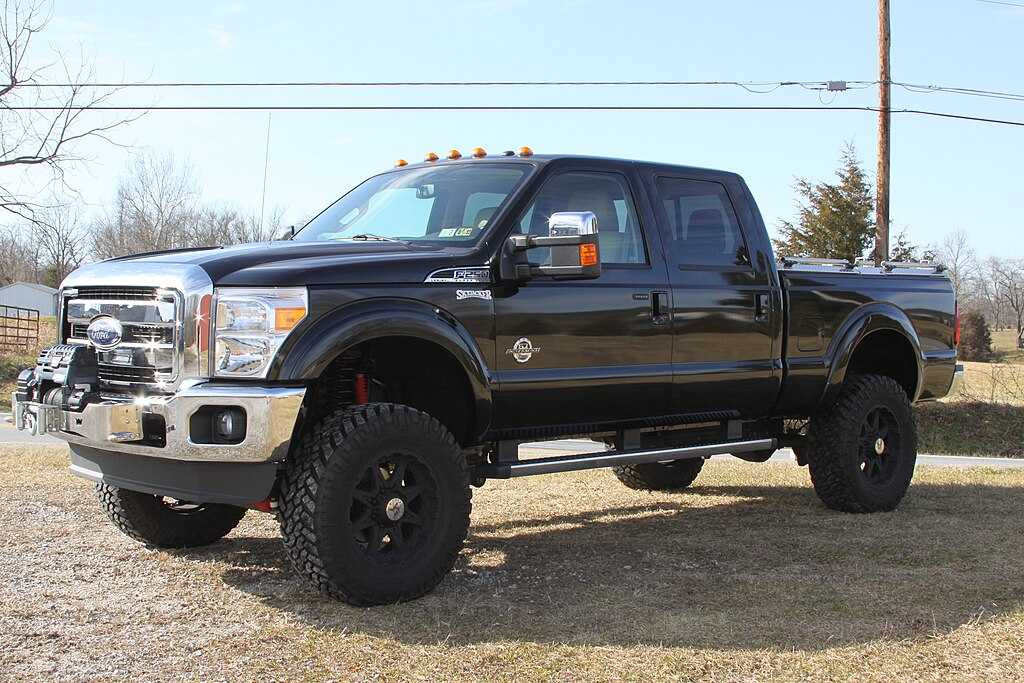 Lifted black Ford F‑250 with large off‑road tires and chrome accents, parked near a wooden fence and trees.