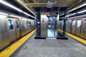 A newly installed glass-enclosed elevator stands between two speeding subway trains on the Queensboro Plaza platform.