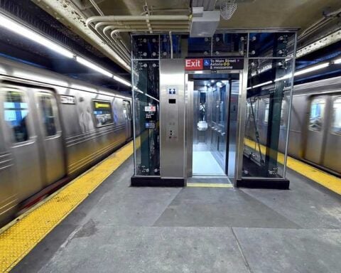 A newly installed glass-enclosed elevator stands between two speeding subway trains on the Queensboro Plaza platform.