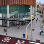 Architectural rendering of the planned 125th Street subway station entrance for the Second Avenue Subway extension, showing a modern glass and metal structure with the Q train visible on an elevated track in the background and pedestrians walking near the entrance.