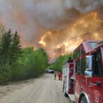 Red fire engine on dirt road with firefighters as a large smoke plume from the Thunderhill Lake Complex wildfire rises near forest in Flin Flon, Manitoba, May 27, 2025