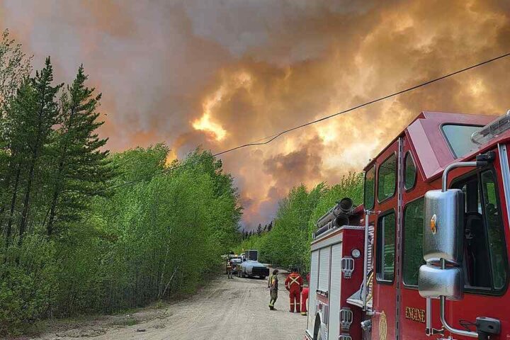 Red fire engine on dirt road with firefighters as a large smoke plume from the Thunderhill Lake Complex wildfire rises near forest in Flin Flon, Manitoba, May 27, 2025