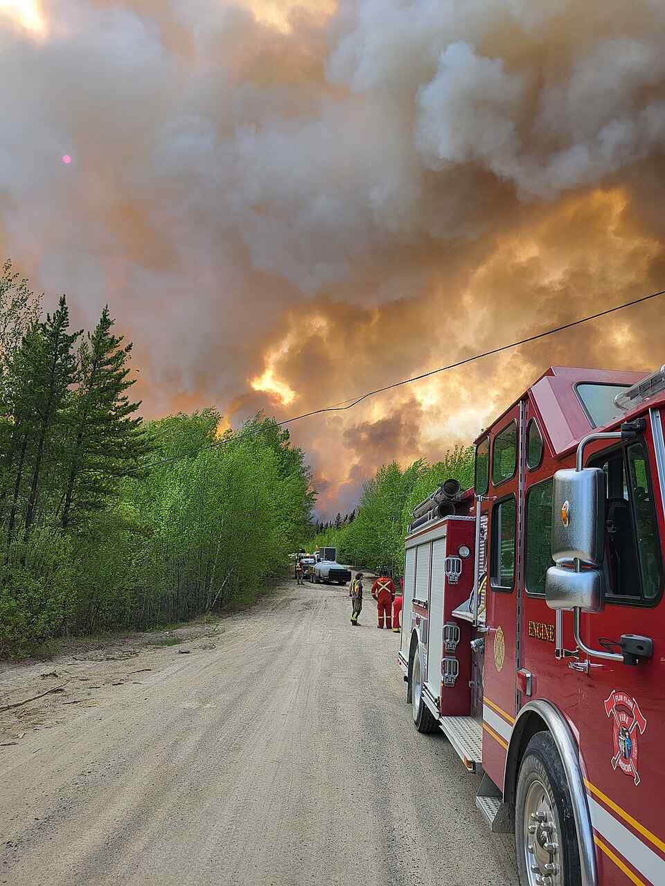 Red fire engine on dirt road with firefighters as a large smoke plume from the Thunderhill Lake Complex wildfire rises near forest in Flin Flon, Manitoba, May 27, 2025