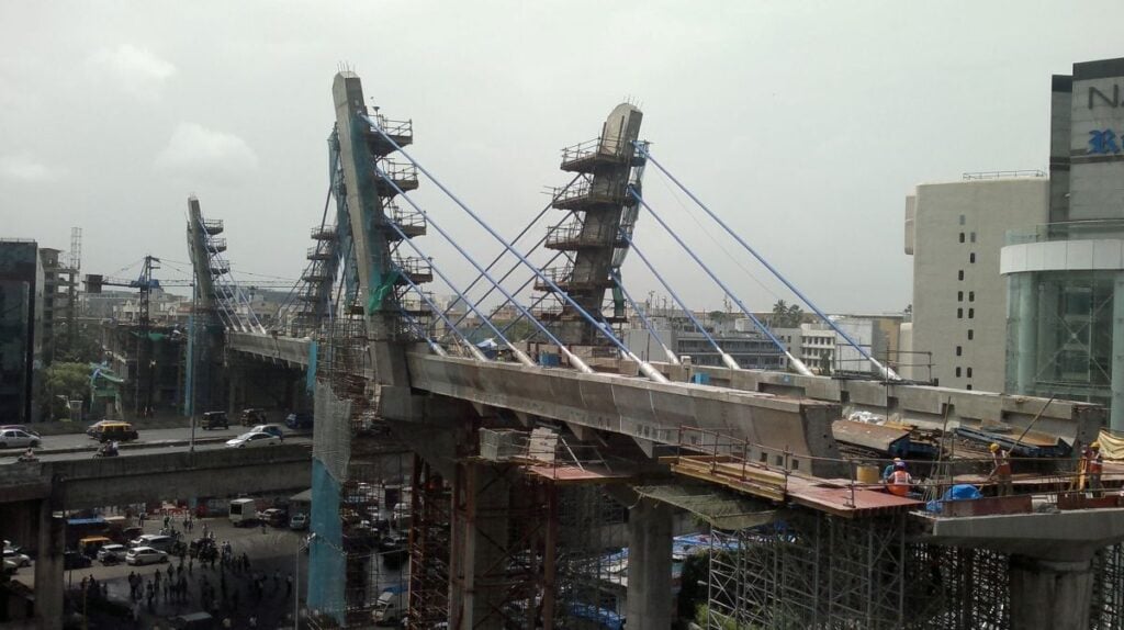 Cable-stayed metro bridge under construction over Mumbai’s Western Express Highway in 2013, with blue stay cables, pylons, scaffolding, workers on deck, and traffic below.