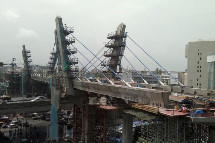 Cable-stayed metro bridge under construction over Mumbai’s Western Express Highway in 2013, with blue stay cables, pylons, scaffolding, workers on deck, and traffic below.