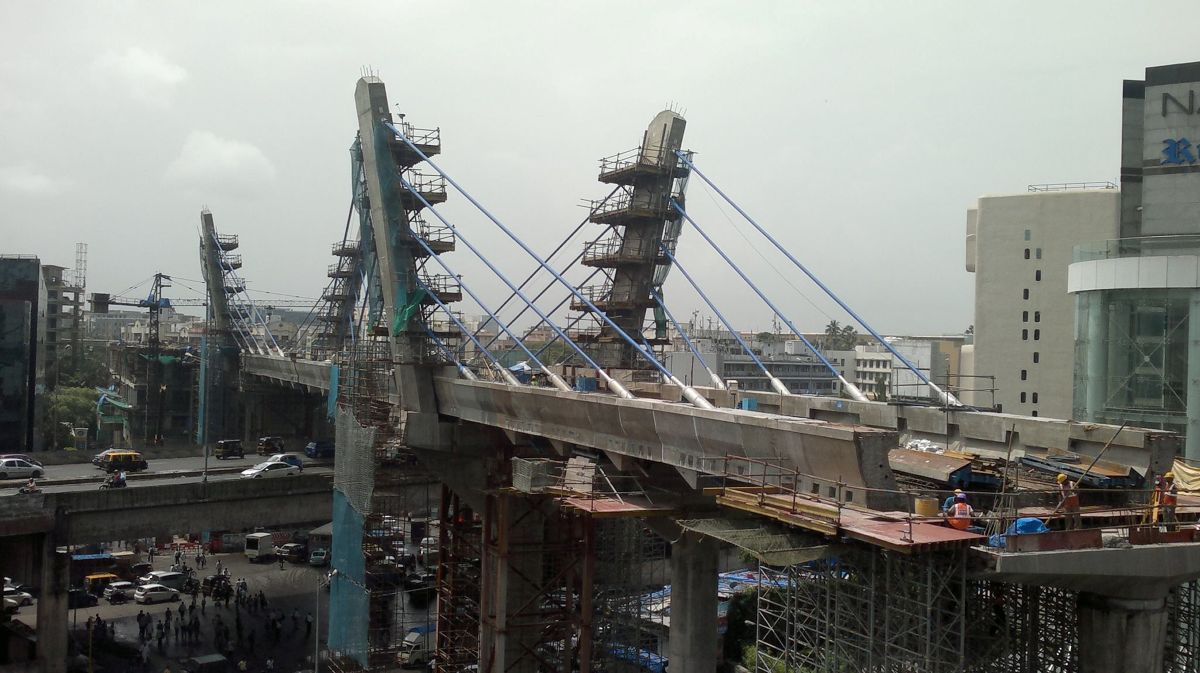 Cable-stayed metro bridge under construction over Mumbai’s Western Express Highway in 2013, with blue stay cables, pylons, scaffolding, workers on deck, and traffic below.