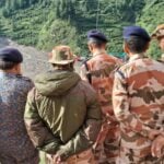 NDRF and ITBP personnel stand on a ridge overlooking a debris-filled valley near Dharali, Uttarkashi; green hills in background and one responder in orange gear at right.
