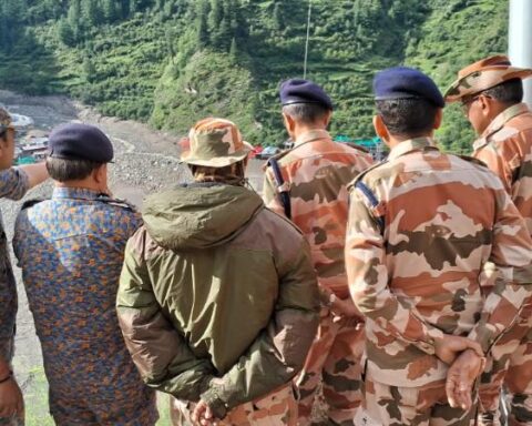 NDRF and ITBP personnel stand on a ridge overlooking a debris-filled valley near Dharali, Uttarkashi; green hills in background and one responder in orange gear at right.