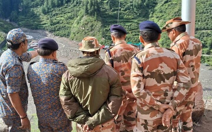 NDRF and ITBP personnel stand on a ridge overlooking a debris-filled valley near Dharali, Uttarkashi; green hills in background and one responder in orange gear at right.