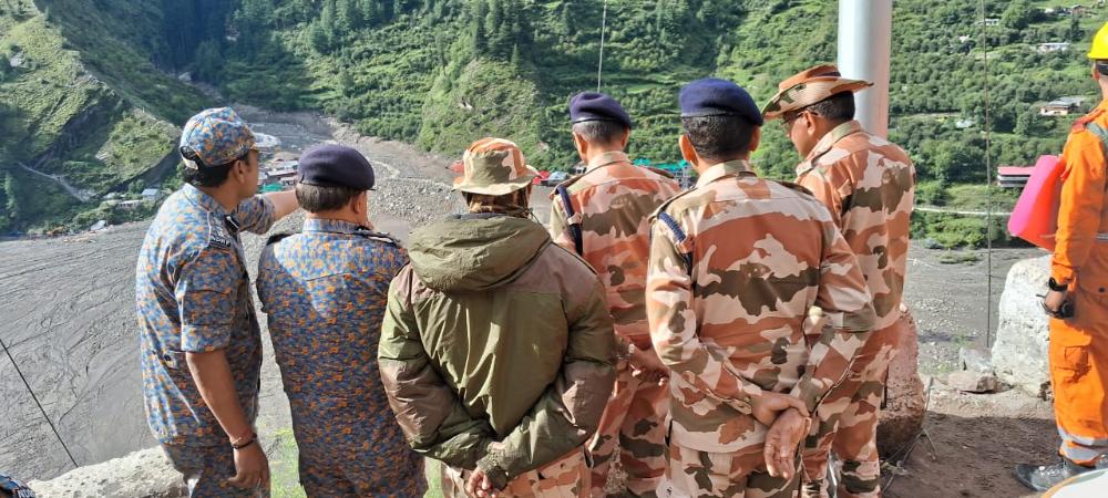 NDRF and ITBP personnel stand on a ridge overlooking a debris-filled valley near Dharali, Uttarkashi; green hills in background and one responder in orange gear at right.