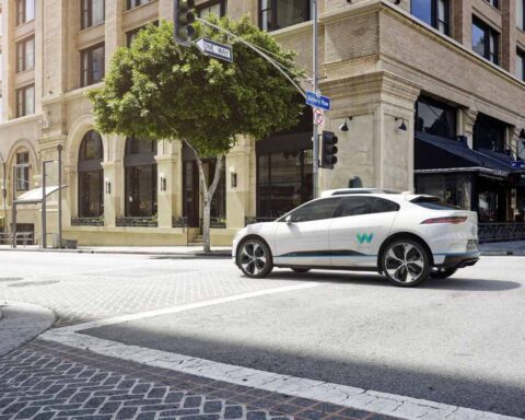 White Jaguar I-PACE Waymo self-driving car with roof sensors turning at a city intersection.