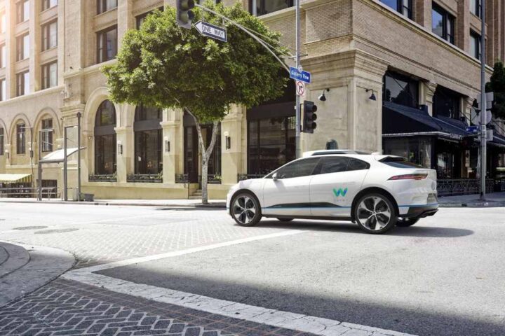 White Jaguar I-PACE Waymo self-driving car with roof sensors turning at a city intersection.