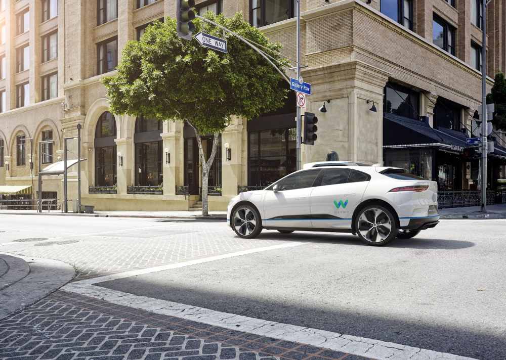 White Jaguar I-PACE Waymo self-driving car with roof sensors turning at a city intersection.