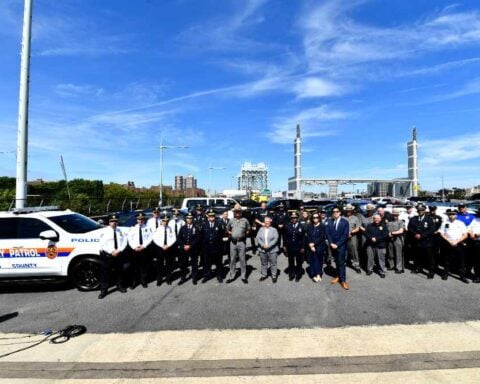 Large group of law enforcement officers from multiple agencies standing together on the RFK Bridge during the 100th ghost plate enforcement operation, with police vehicles and the bridge structure visible in the background.