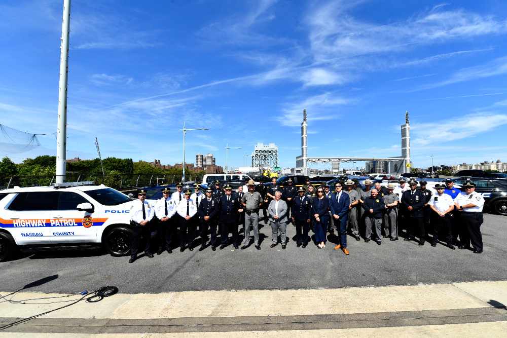 Large group of law enforcement officers from multiple agencies standing together on the RFK Bridge during the 100th ghost plate enforcement operation, with police vehicles and the bridge structure visible in the background.
