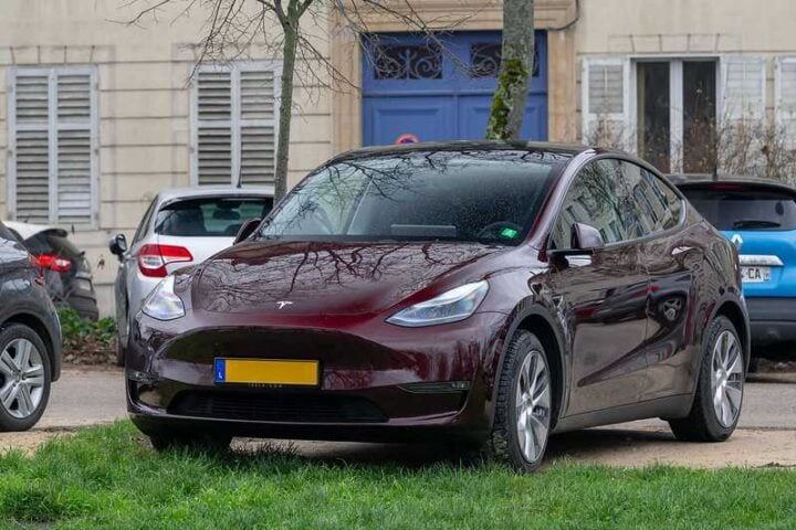 Maroon Tesla Model Y parked on grass beside a sidewalk in front of a beige building with shutters in Nancy, France; yellow EU plate; nearby cars.