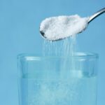 Spoon pouring granulated sweetener into a glass of water against a blue background.