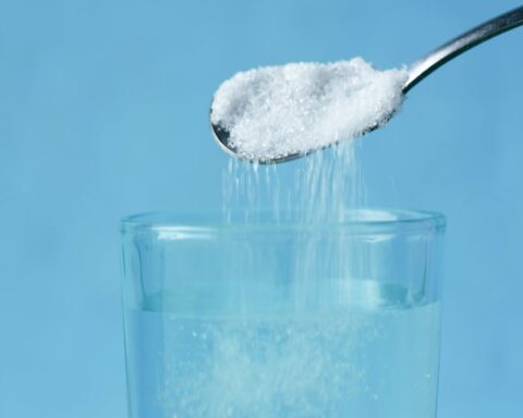 Spoon pouring granulated sweetener into a glass of water against a blue background.