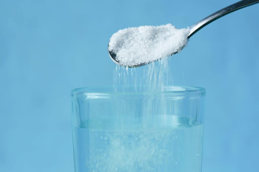 Spoon pouring granulated sweetener into a glass of water against a blue background.