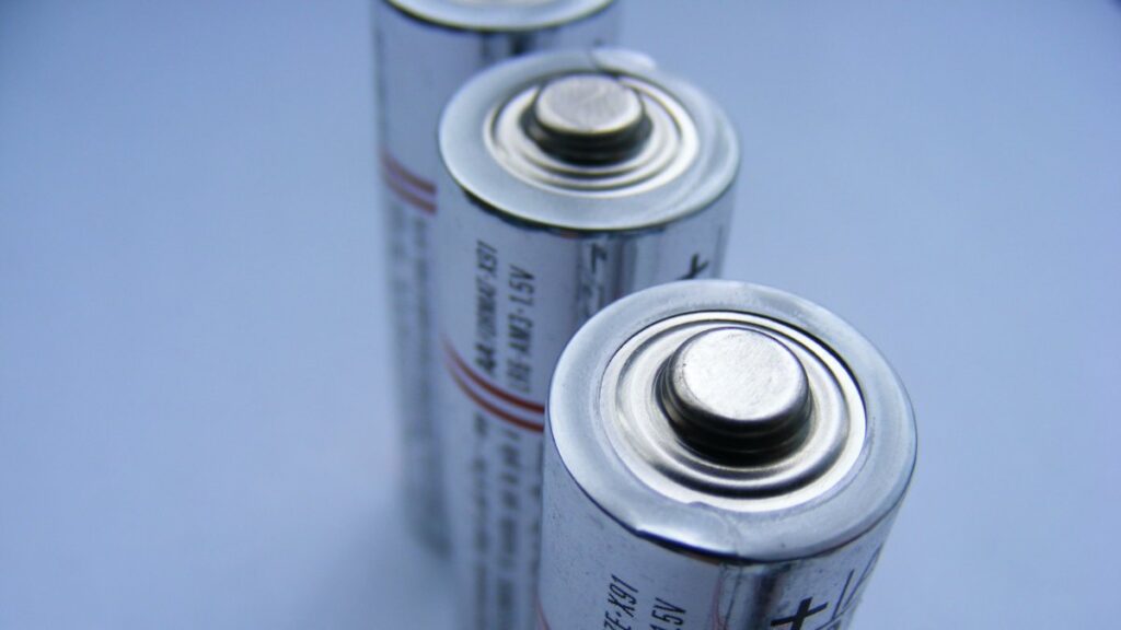 Close-up photograph of two cylindrical batteries standing vertically against a blue background, showing their metal positive terminals.
