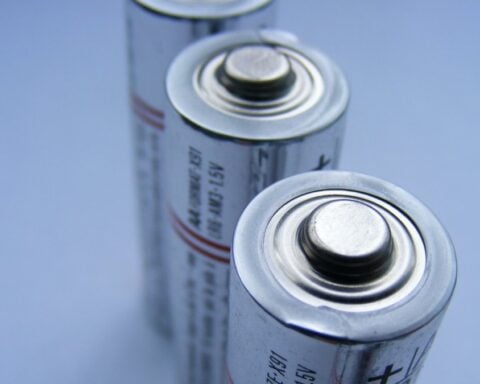 Close-up photograph of two cylindrical batteries standing vertically against a blue background, showing their metal positive terminals.