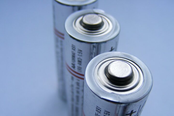Close-up photograph of two cylindrical batteries standing vertically against a blue background, showing their metal positive terminals.