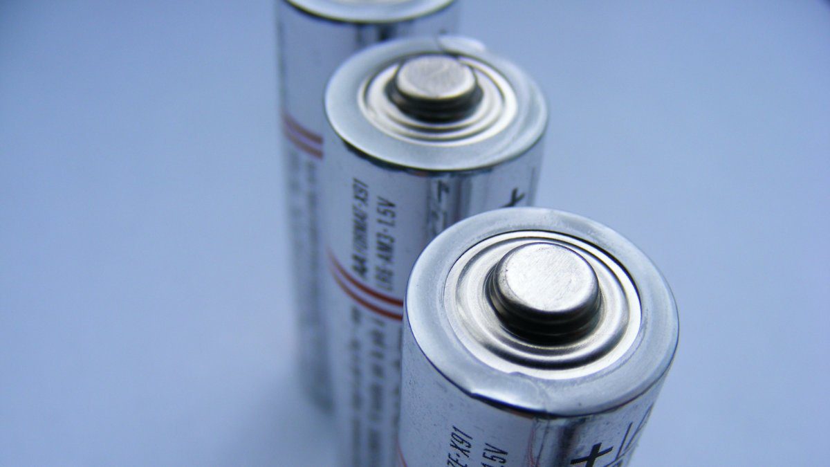 Close-up photograph of two cylindrical batteries standing vertically against a blue background, showing their metal positive terminals.