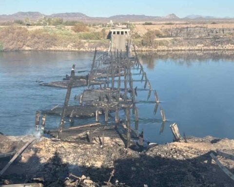 A burned-out wooden bridge, its skeletal remains jutting across a calm river, surrounded by scorched terrain and distant scrubland.